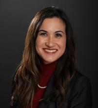 smiling woman with long brown hair wearing a maroon turtleneck shirt, pearl necklace and black jacket