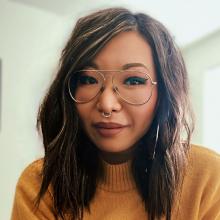 woman with medium length wavy dark hair and light highlights, wearing silver-framed glasses, nose ring, hoop earrings and yellow-orange sweater