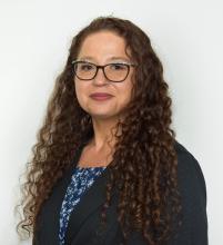 woman with long curly auburn hair and glasses, wearing a dark jacket over blue patterned shirt