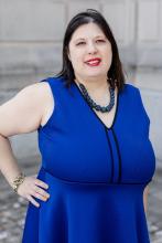 smiling woman with medium length dark hair, wearing a blue dress and teal necklace