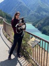 Woman in windy outdoor setting in front of mountains and a lake. She is holding a small black, white, and tan dog.