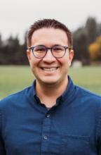 Mixed-race Chinese and Caucasian man with dark brown hair, blue glasses, and a blue button-down shirt smiling while standing in a field.