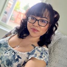 Asian woman with wavy, black, shoulder length hair, wearing glasses and a white blouse with blue flowers